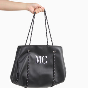 Bolso grande Xava negro de material sintético con textura perforada, iniciales bordadas y tirantes de cuerda gráfica.